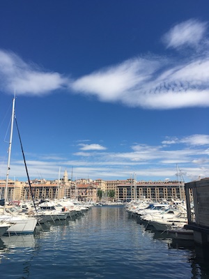 Several boats lined up in the port of Marseille with the city in the background and a wide blue sky with a few clouds.
